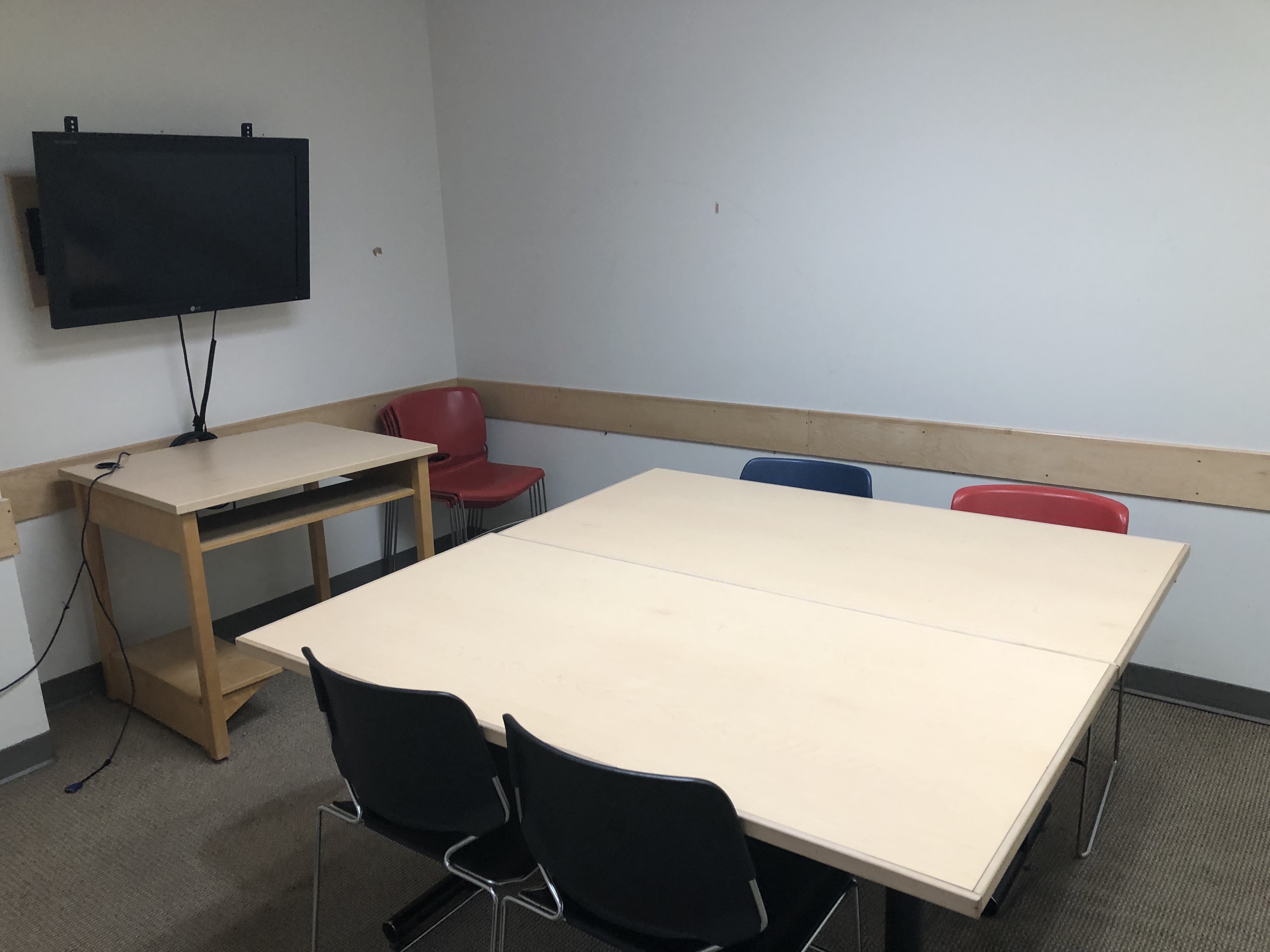 Photo of group study room. Rooms described below.  The image shows two tables put together to create a square. Four chairs at desk and four chairs stacked in corner. 