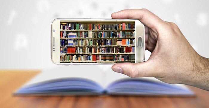A hand holding a cell phone showing a library shelf. The hand is overtop an open textbook.