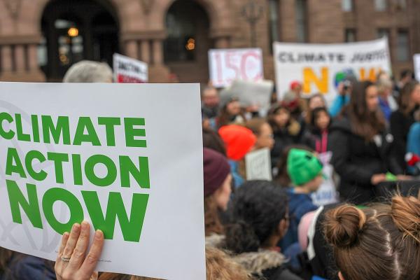 A hand holding a sign that reads ‘Climate Action Now’ in front of a large group of people. Climate justice related signs are in the background.
