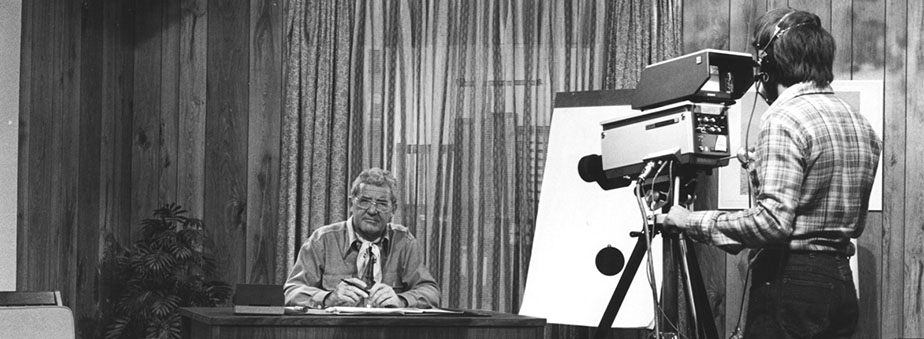 Man sitting at desk in front of a camera and cameraman.