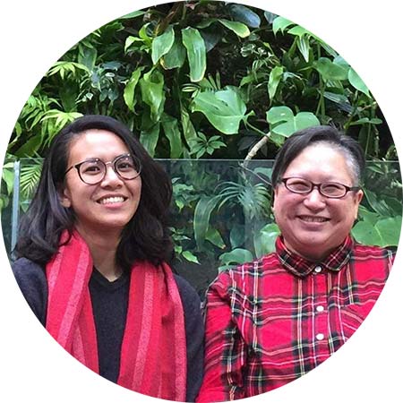 Artists Althea Balmes (she/her) and Jo SiMalaya Alcampo (they/them, siya), known together a Kwentong Bayan Collective, are smiling at the camera sitting side by side. On the left, Althea is wearing a red scarf that matches Jo’s red shirt on the right. The artists are pictured in front of a background of lush green plant leaves. The picture is cropped into a circle.