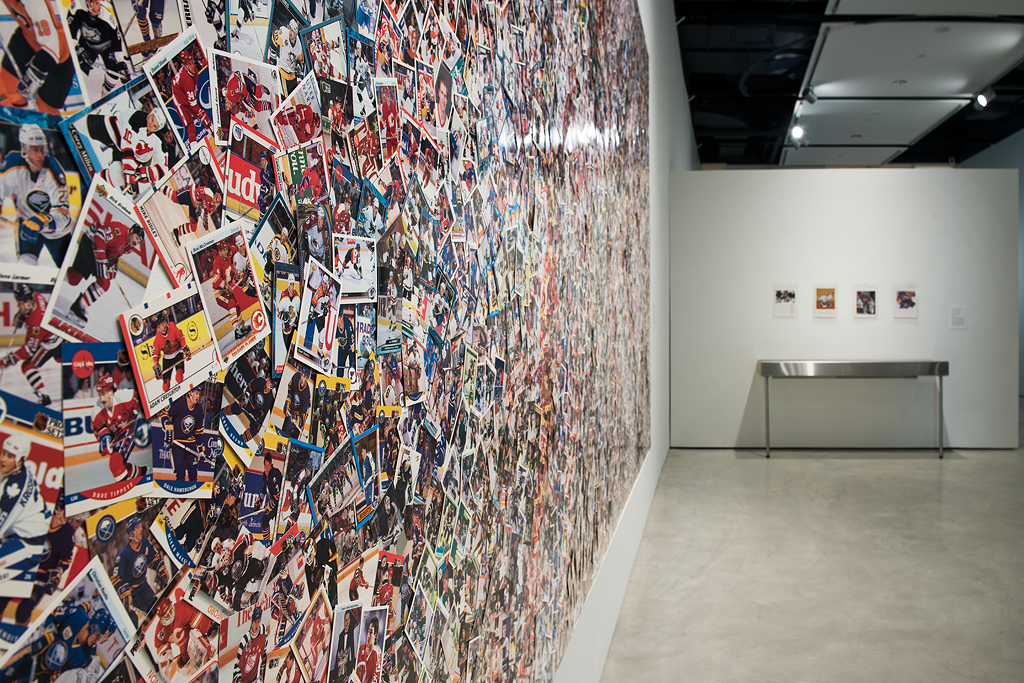 Installation photo of collage from an angled perspective. In the foreground, the overlapping hockey cards are visible with predominantly red, yellow, blue, and white colours. The perspective of the image fades toward the background.