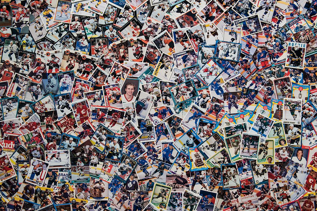 A detail of the large abstract collage fills the frame showcasing hundreds of hockey cards. Here, the cards can be seen as having been laid in a subtle pattern of concentric circles starting from the centre of the image. The cards overlap haphazardly, some images on angles, some upside down. A few images on the cards stick out randomly, like a close-up of a hockey player’s face near the centre of the collage, a player celebrating a win near the left of the collage, and a goalie near the right of the collage.