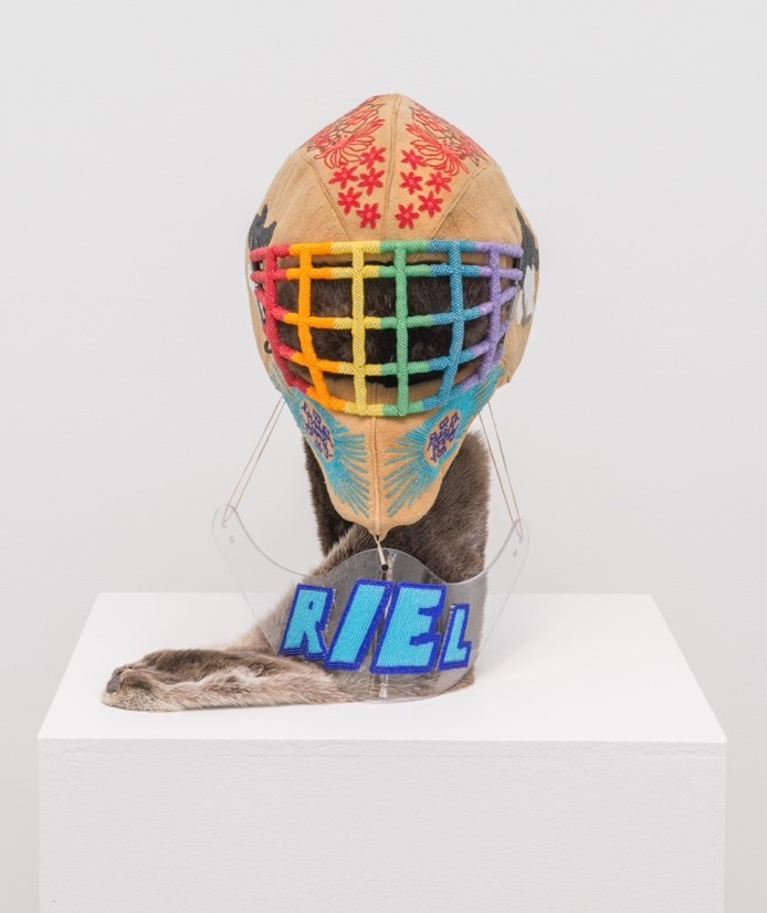 An otter pelt extends down from a goalie mask and lays on a white plinth. Overtop is a clear Plexi glass neck guard with blue beaded letters that spell the name “Riel”. Above the pelt is an otter skin goalie mask with Indigenous flag imagery and red maple leaves. The face cage of the mask is beaded in rainbow colours. Two images of this sculpture are presented, showing a front facing view and an oblique angled view of the work
