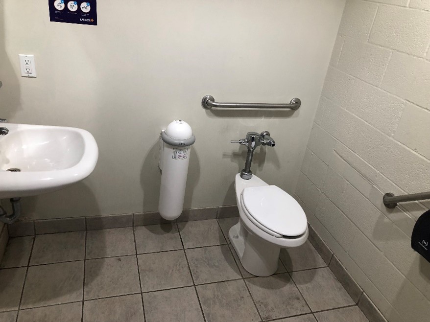 The interior of the individual use washroom. It has a sink at 11 o’clock, 12 o’clock is a menstrual hygiene disposal unit mounted to the wall. It has a domed top and if you hover hand over top, the dome will lift revealing a space to dispose of hygiene products. Toilet is at 1 o’clock.