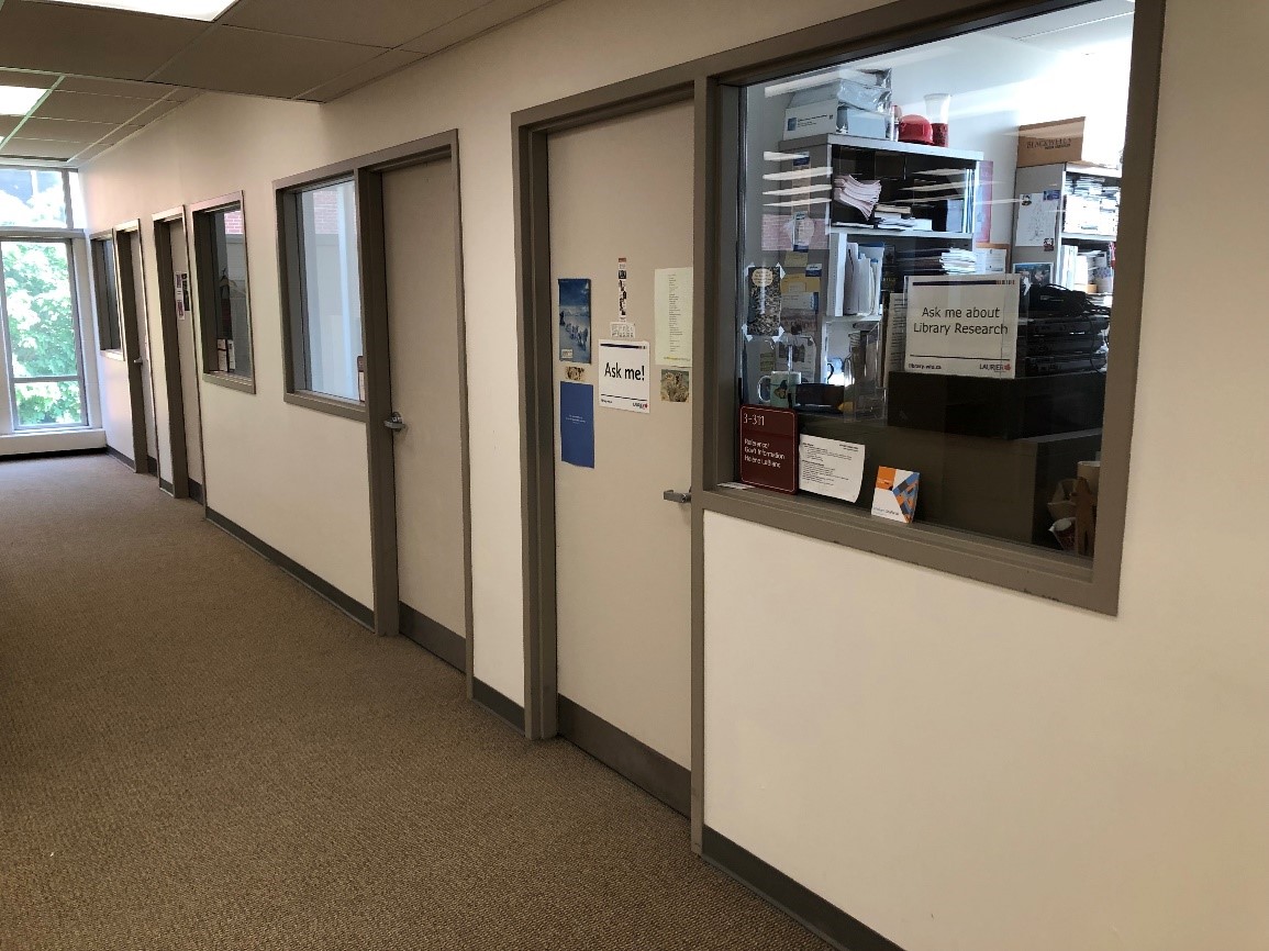 A series of office doors for librarians. The rooms also have windows into them. One door and window has signs on the outside for students, saying ‘Ask me’ and ‘Ask me about Library Research’