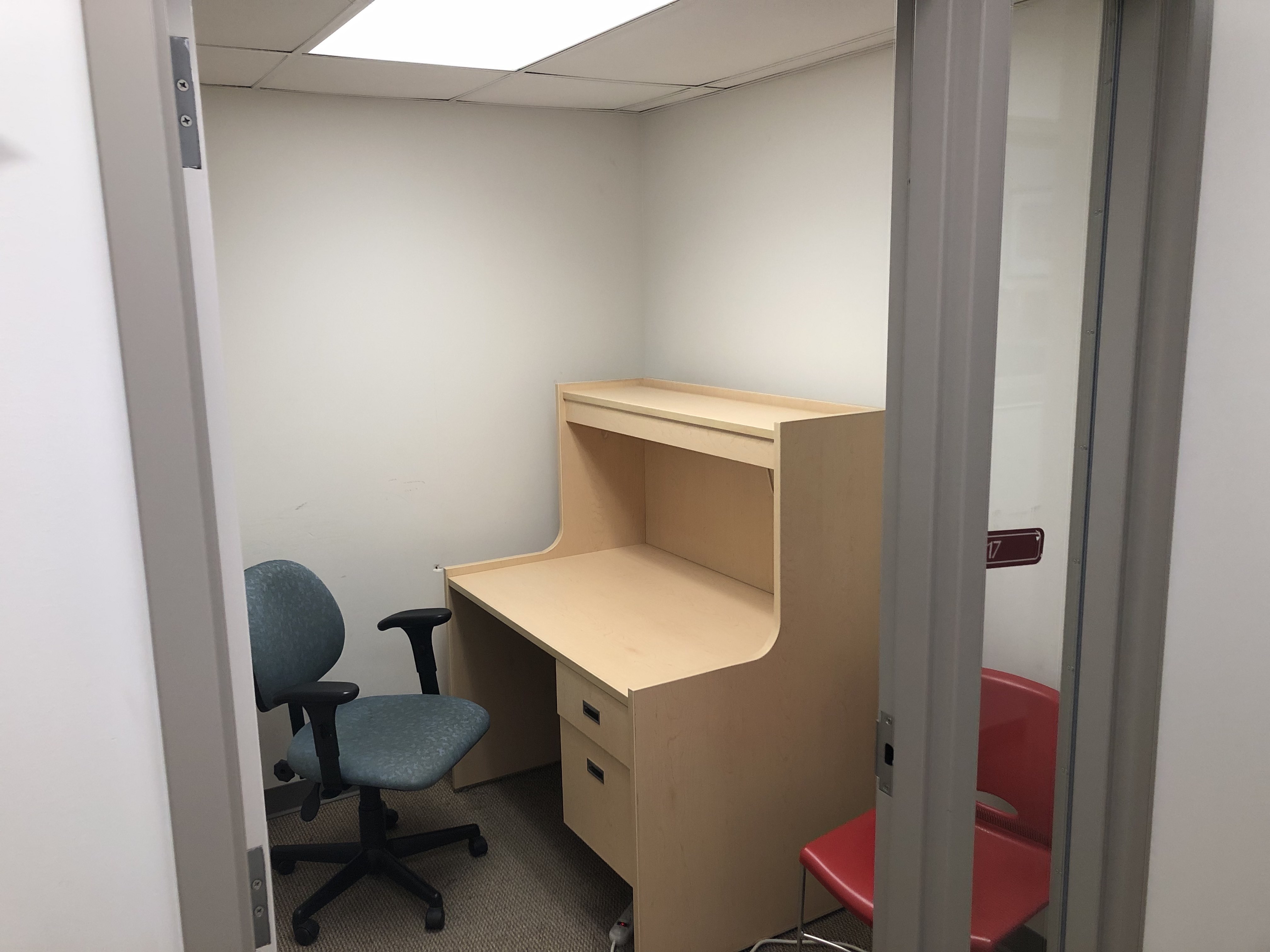 A private study room with a chair and desk. The desk has draws. There is a tall, narrow window next to door