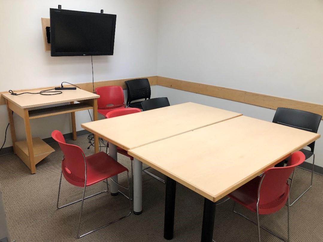 A group study room with two tables pressed together to create a large table. Moveable chairs are around it with a few more chairs against of wall. A monitor is mounted on the wall with a cable onto a table. There is a window from the study room into the hallway