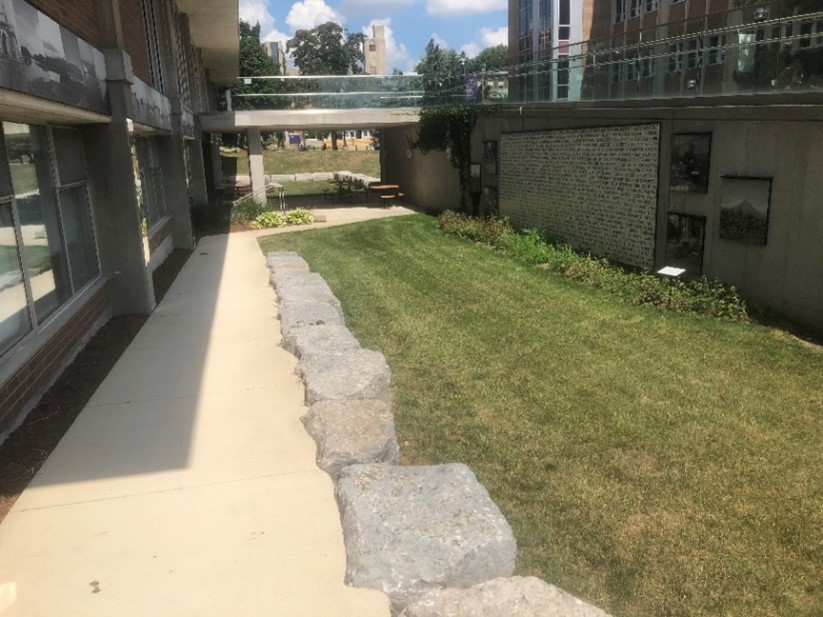 The sunken garden. A concrete path with large irregular stones on the right. It gently descends into the sunken garden area. Following the path leads to some fixed picnic tables. Crossing above the tables is the walkway from the ramp to the front doors. Visual art is along walls of the sunken garden, emphasizing Indigenous plants