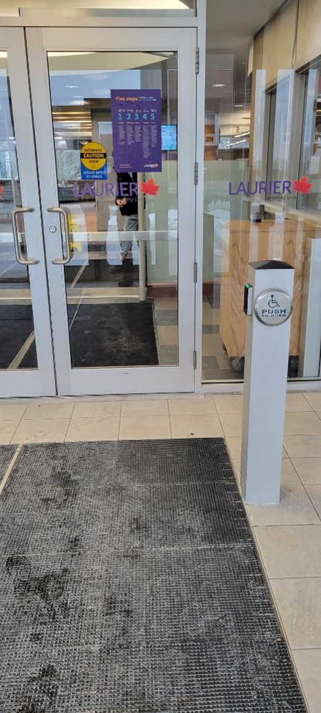 Between the exterior doors and interior library doors. A rectangular bollard mounted with a round button to open the interior library doors. On another side of the bollard is smaller rectangular to tap your OneCard in situations where the library is open but the building locked, for example during homecoming weekend.