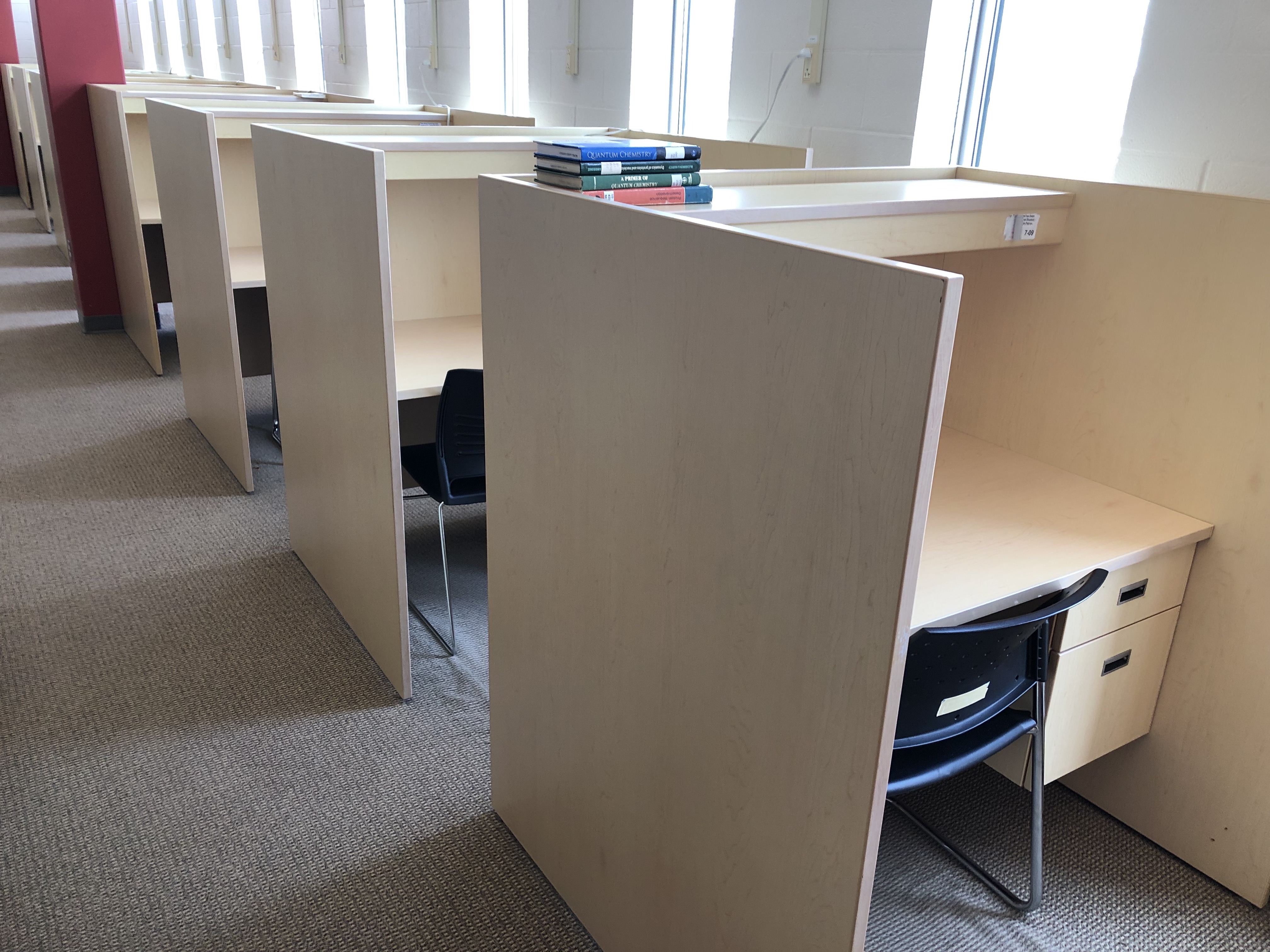 A line of study carrels along the outer wall of the library. These desks have two drawers on the right side. There are partitions on the left and right side of these desks, however, these desks are arranged face front to back with other desk. They are still separated by a partition for privacy.