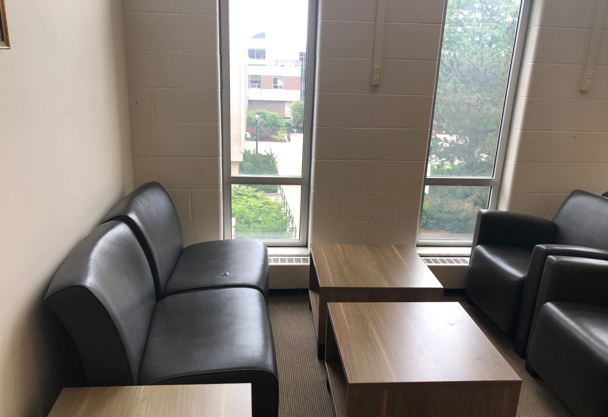 Comfy chairs next to each other some with arm rests, some without. View of window. Shin height coffee tables. Wall mounted outlet on wall between windows, approximate 4 feet above floor