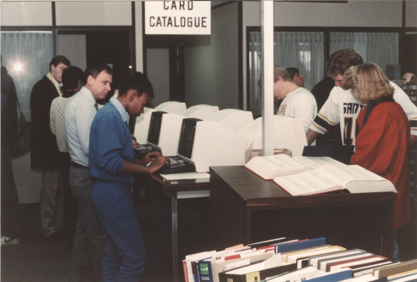 Librarian and students gathered around computer terminals with books on a cart in the foreground