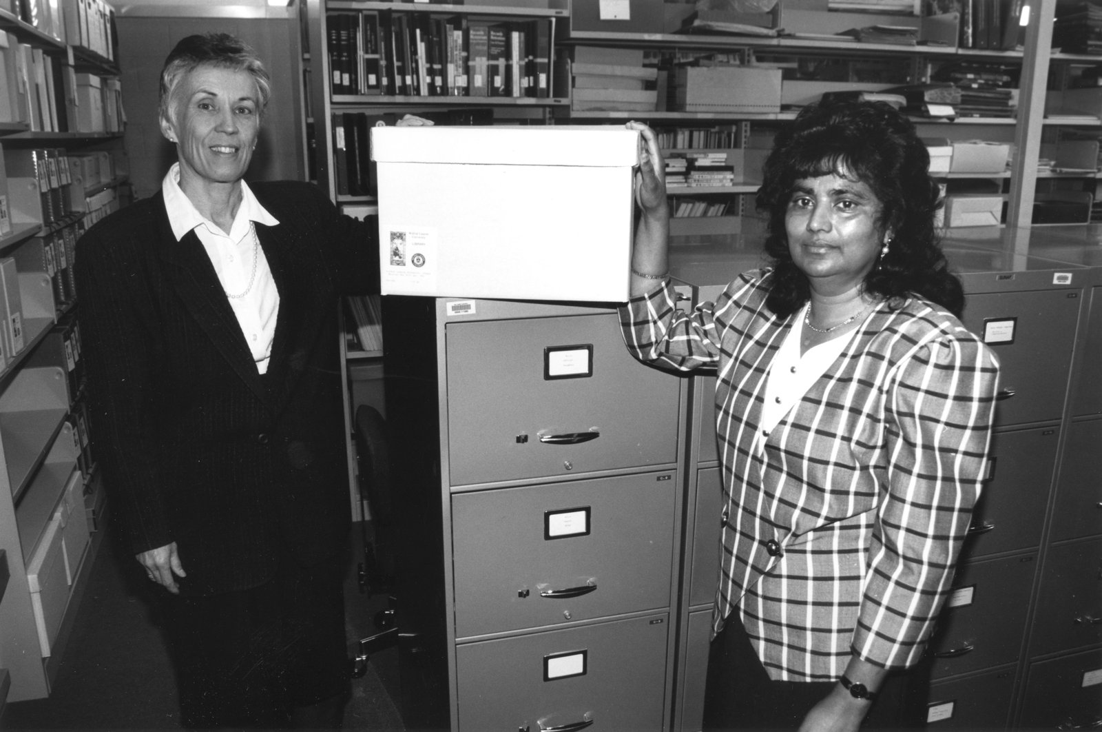 Two people standing with a box on top of filing cabinets with shelves of boxes and books in the background