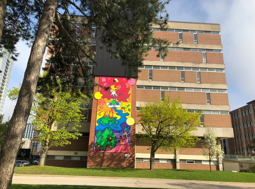 South wall of library. Between two maple trees is a mural made of 18 panels. At base, elder and child hand 3 bear cubs blueberries. At the top is an eagle and 4-part medicine wheel
