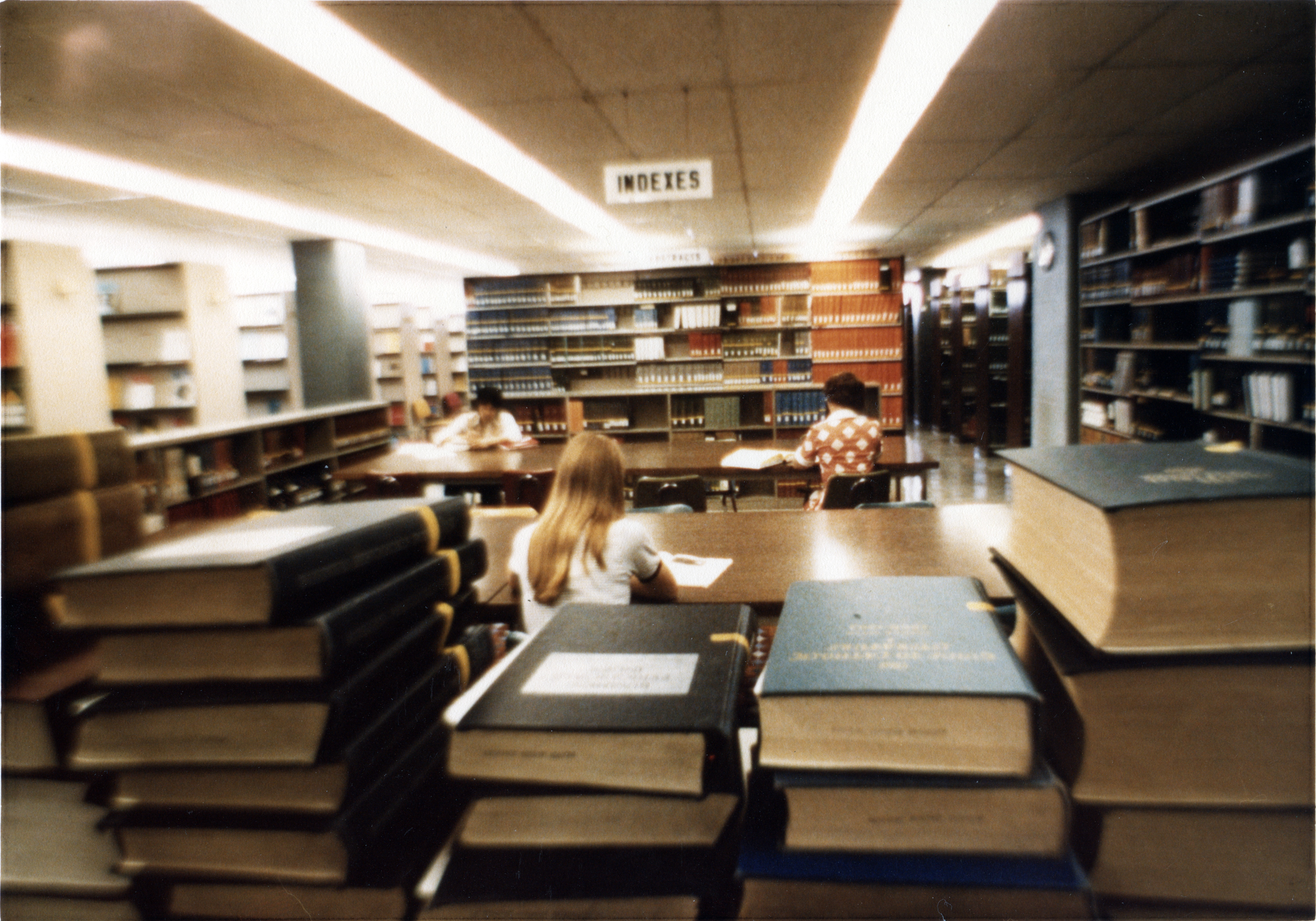 Students studying at desks with stacks of books in the foreground and bookshelves in the background