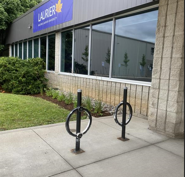 Two black, circular bike racks positioned on concrete close to the entrance of the building. Behind them is some grassy area and large windows into the building.