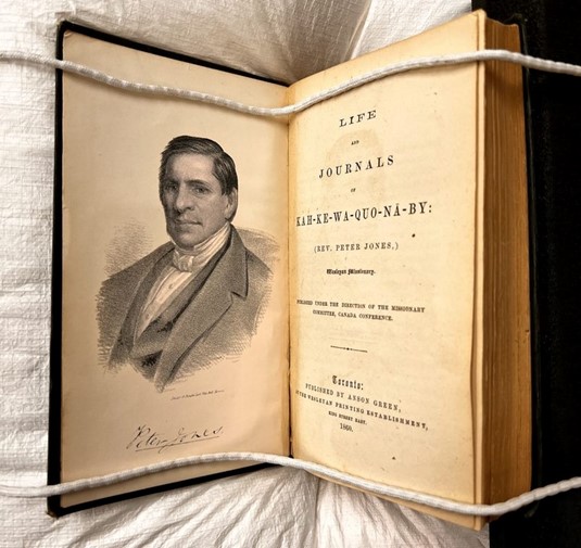 Book opened on a book cover with snake weights holding the pages in place. Left page is an image of Peter Jones and the right page is the title page. Book is Life and Journals of Kah-ke-wa-quo-nā-by.