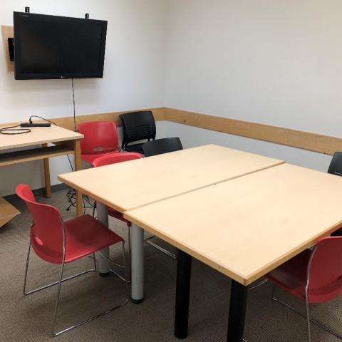 Group study room with two tables pushed together. Some chairs are stacked in a corner next to a second table to hold laptops to connect to a monitor on the wall. Three chairs are around the main table.