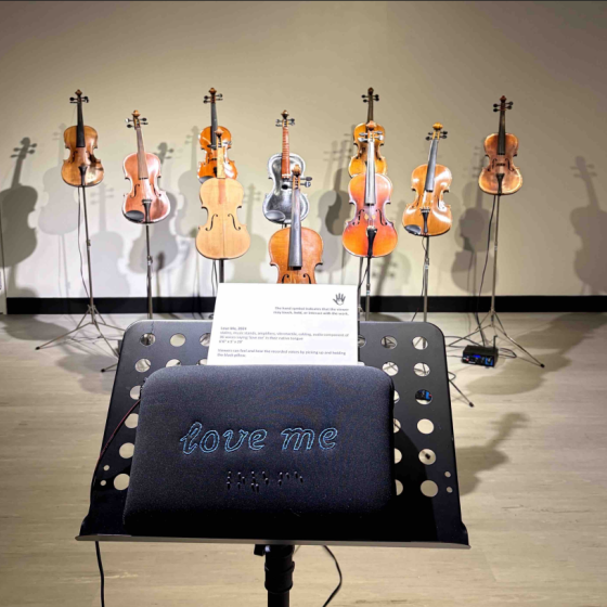A music stand holds a small pillow with the words in script and braille, "love me". By holding the pillow, viewers can feel and hear recorded voices. In the background are seven violins that are part of an integrated audio component