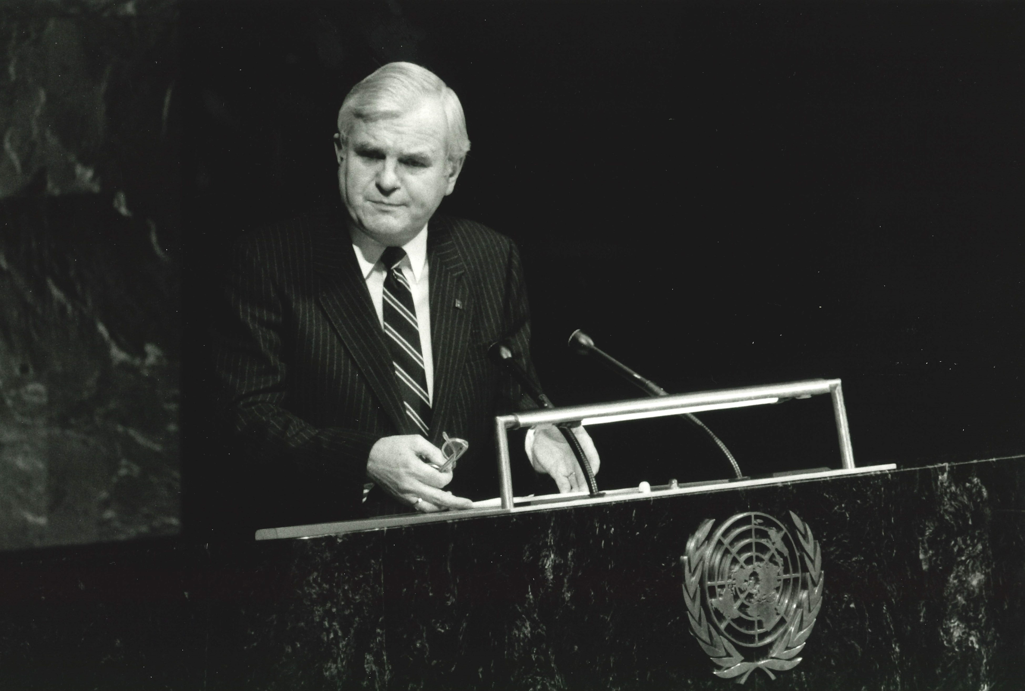 A black-and-white photograph of Walter McLean giving a speech at a podium with the United Nations emblem on it. Walter has light toned skin and hair and is wearing a dark suit and striped tie.